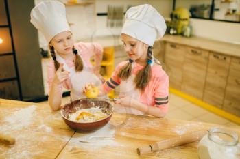 Two little girls cooks in caps rubs lemon to the bowl, cookies preparation on the kitchen. Kids cooking pastry, children chefs preparing cake