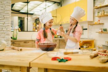 Two little girls cooks in caps tastes sweet vanilla powder, cookies preparation on the kitchen. Kids cooking pastry, children chefs preparing cake