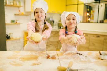 Two smiling little girls cooks in caps shows dough balls, cookies preparation on the kitchen. Kids cooking pastry, children chefs preparing cake