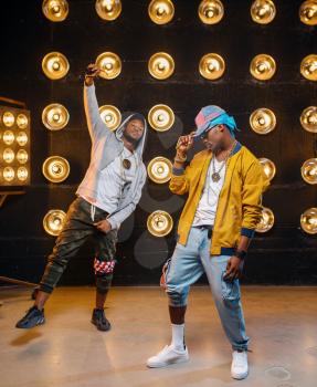 Two black rappers in caps, dance perfomance in club, stage with spotlights on background. Rap performers on scene with lights, underground music concert, urban style
