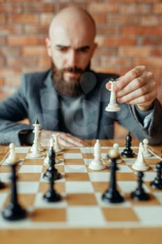 Male chess player playing white figures, queen move. Chessplayer at board, front view, intellectual tournament indoors. Chessboard on wooden table, strategy game