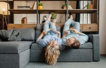 Young love couple with books lying on the couch upside down. Husband and wife relax in living room. Happy family having fun together