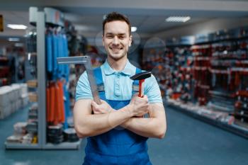 Smiling male worker in uniform holds hammer and corner in tool store. Choice of professional equipment in hardware shop, instrument supermarket