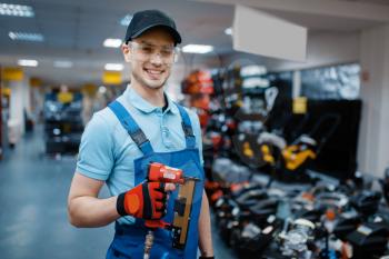 Male worker in uniform holds pneumatic nailer in tool store. Choice of professional equipment in hardware shop, instrument supermarket