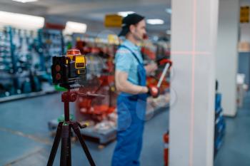 Male worker in uniform testing laser level on tripod in tool store. Choice of professional equipment in hardware shop, instrument supermarket