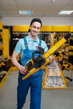 Male worker in uniform holds chainsaw in tool store. Choice of professional equipment in hardware shop, electrical instrument supermarket