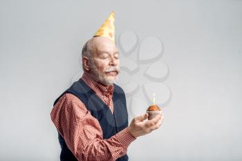 Smiling elderly man in party cap shows cake with candle, grey background. Cheerful mature senior looking at camera in studio