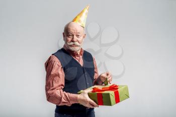 Happy elderly man in party cap holds gift box, grey background. Cheerful mature senior looking at camera in studio
