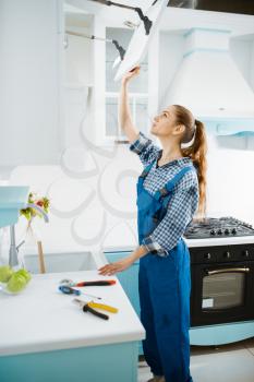 Cute female furniture maker in uniform repair cupboard in the kitchen. Handywoman fixing problem with garniture cabinet, repairing service at home
