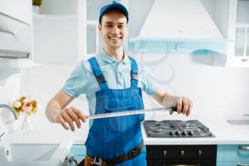 Smiling male furniture maker in uniform holds measuring tape in the kitchen. Handyman installing garniture, repairing service at home