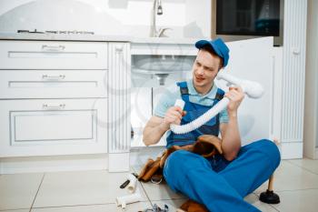 Male plumber in uniform listerns to the drain pipe in the kitchen, humor. Handywoman with toolbag repair sink, sanitary equipment service at home