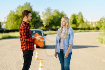 Male instructor with traffic cone and student on road, lesson in driving school. Man teaching lady to drive vehicle. Driver's license education