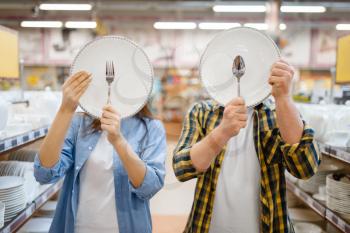 Young couple jokes with plates in houseware store. Man and woman buying home goods in market, family in kitchenware supply shop