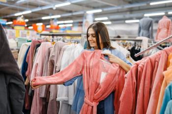 Young woman choosing bathrobe in bed linen store. Female person buying home goods in market, lady in bedding shop