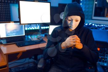 Hacker in mask and hood sitting at his workplace with laptops and PC, information hacking. Internet spy, illegal lifestyle, risk job