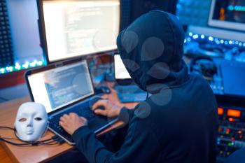 Hacker in mask and hood sitting at his workplace with laptop and PC, password or account hacking. Internet spy, illegal lifestyle, risk job, network criminal