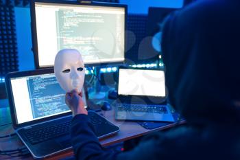 Hacker in hood holds mask in hand and at his workplace with laptop and PC, password or account hacking. Internet spy, crime lifestyle, risk job, network criminal