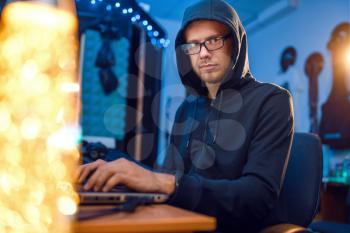 Hacker in hood at his workplace with laptop and desktop PC, website or corporate hacking, darknet user. Internet spy, crime lifestyle, risk job, network criminal