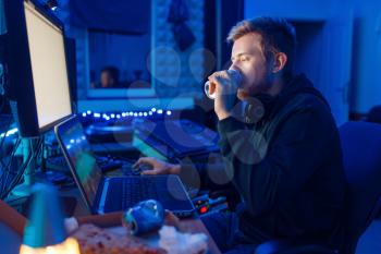 Male gamer drinking an energy drink at his workplace with laptop and desktop PC, gaming night lifestyle. Computer games player in his room with neon light, streamer