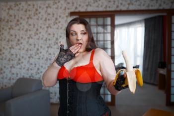 Overweight woman in erotic lingerie amazement looks at the banana. Sexy overweight girl with big breast, corrupt large size lady