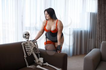 Overweight woman in erotic lingerie and human skeleton are sits on couch. Sexy overweight girl with big breast, corrupt large size lady