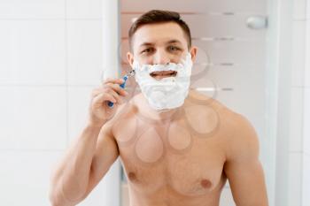 Man shaves his beard with razor in bathroom, routine morning hygiene. Male person performs skin and body treatment procedures