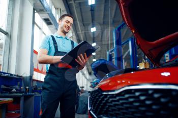 Worker with a checklist stands at vehicle with opened hood, car service station. Automobile checking and inspection, professional diagnostics and repair