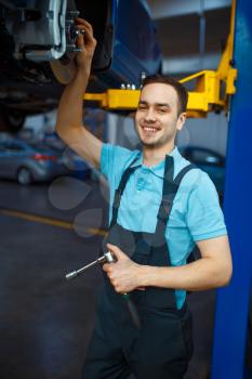 Repairman in uniform repairing vehicle on lift, car service station. Automobile checking and inspection, professional diagnostics and repair