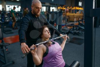 Instructor helps to overweight woman on exercise machine, gym. Female person struggles with excess weight, aerobic workout against obesity