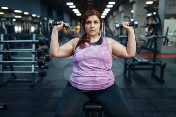 Overweight woman poses with dumbbells in gym, active training. Obese female person struggles with excess weight, aerobic workout against obesity, sport club
