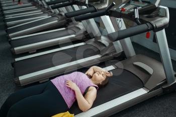 Tired overweight woman lying on treadmill in gym, leisure after active training. Obese female person struggles with excess weight, aerobic workout against obesity, sport club
