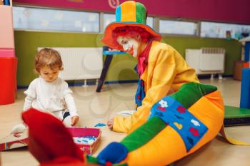 Funny clown play with joyful little girl together. Birthday celebrating in playroom, baby holiday in playground. Childhood happiness, childish leisure