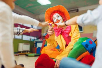 Funny clown gives out lollipops to happy children. Birthday party celebrating in playroom, baby holiday in playground. Childhood happiness, childish leisure