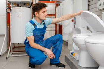 Plumber in uniform choosing the toilet at showcase in plumbering store. Man with notebook buying sanitary engineering in shop