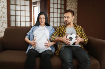 Husband with ball and his pregnant wife with belly joking at home, humor. Pregnancy, prenatal period. Expectant mom and dad are resting on sofa, health care