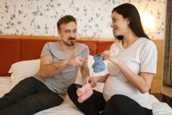 Happy couple, pregnant wife with belly shows to husband clothing for newborns at home, bedroom interior on background. Pregnancy, prenatal period. Expectant mom and dad are resting in bed