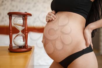 Pregnant woman with belly poses against sandglass at home, side view. Pregnancy, calm in prenatal period. Expectant mom resting in bedroom, healthy lifestyle