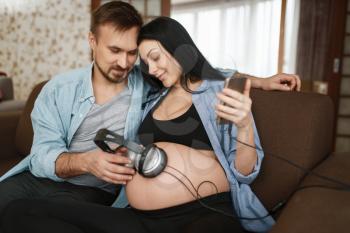 Husband and pregnant wife with headphones at her belly. Pregnancy, prenatal period at home. Expectant mom and dad are resting on sofa, health care