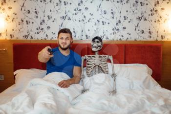 Man and human skeleton watching TV in the bed, bad relationship. Couple having a problems, family quarrel or conflict