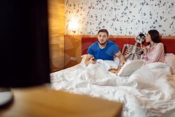 Husband watching TV, wife kissing human skeleton in the bed, bad relationship. Couple having a problems, family quarrel, conflict of married man and woman