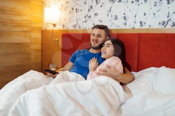 Love couple in pajamas lying in bed and watching TV at home, good morning. Harmonious relationship in young family. Man and woman resting together in their house, carefree weekend