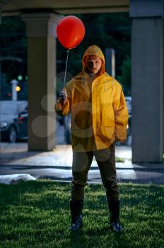 Scary man with balloon walking in night summer park in rainy day. Strange male person in rain cape and rubber boots, wet weather in alley