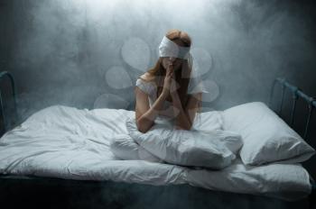 Blindfolded crazy woman sitting in bed, dark room on background. Psychedelic female person having problems every night, depression and stress, sadness, psychiatry hospital