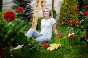 Smiling woman in headphones works with flowers in the garden. Female gardener takes care of plants outdoor, gardening hobby, florist lifestyle and leisure