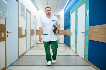 Friendly male doctor in uniform holds notebook in clinic corridor. Professional medical specialist in hospital, laryngologist or otolaryngologist, gynecologist or mammologist, surgeon