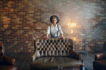 Woman in strict clothes and hat poses in studio, retro fashion, gangster style. Vintage business lady in office with brick walls