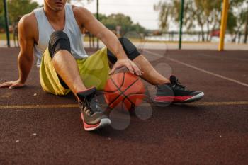 Basketball player with ball sitting on the ground on outdoor court. Male athlete in sportswear resting after streetball training