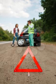 Emergency stop sign, car breakdown, couple calling for tow truck on background. Broken automobile or repairing of flat tyre on vehicle, trouble with punctured auto tire on highway