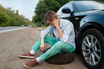 Car breakdown, young man sitting on spare tyre. Broken automobile or problem with vehicle, trouble with punctured auto tire on highway