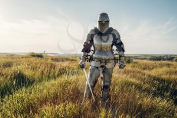 Knight in armor and helmet holds sword. Armored ancient warrior in armour posing in the field